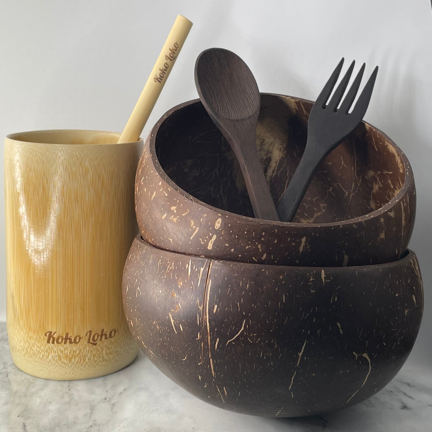 Koko Loko Starter Collection featuring two natural coconut shell bowls stacked with an ebony wood spoon and fork inside, alongside a Koko Loko engraved bamboo cup with a bamboo straw on a marble surface, eco-friendly sustainable gift set by Koko Loko