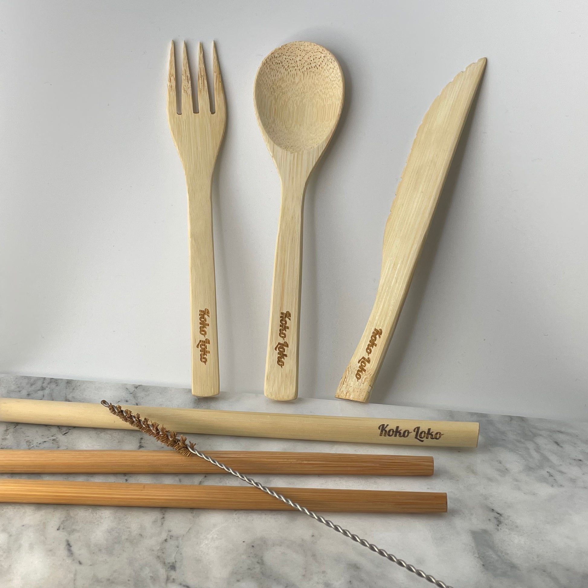 Panda Cutlery Collection handcrafted bamboo fork, spoon and knife with Koko Loko engraving standing upright against a white wall above bamboo chopsticks, straw and cleaner brush on a marble surface, eco-friendly sustainable cutlery set by Koko Loko
