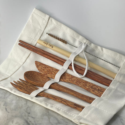 Earth Cutlery Collection coconut wood fork, spoon and knife with dark wood chopsticks, bamboo straw and cleaner brush laid out in an open white cotton roll pouch with Koko Loko engraving on a marble surface, eco-friendly sustainable cutlery set by Koko Loko
