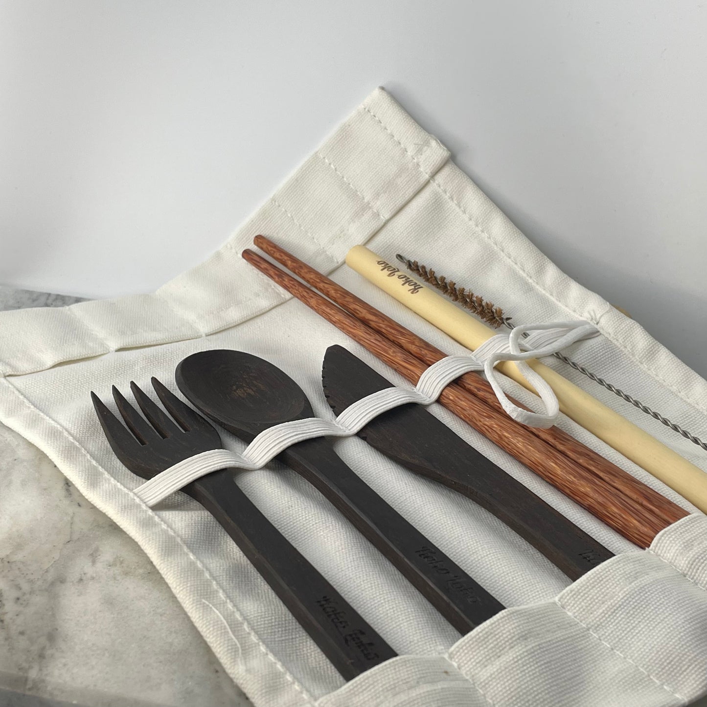 Nirvana Cutlery Collection ebony wood fork, spoon and knife with dark wood chopsticks, bamboo straw and cleaner brush laid out in an open white cotton roll pouch with Koko Loko engraving on a marble surface, eco-friendly sustainable cutlery set by Koko Loko
