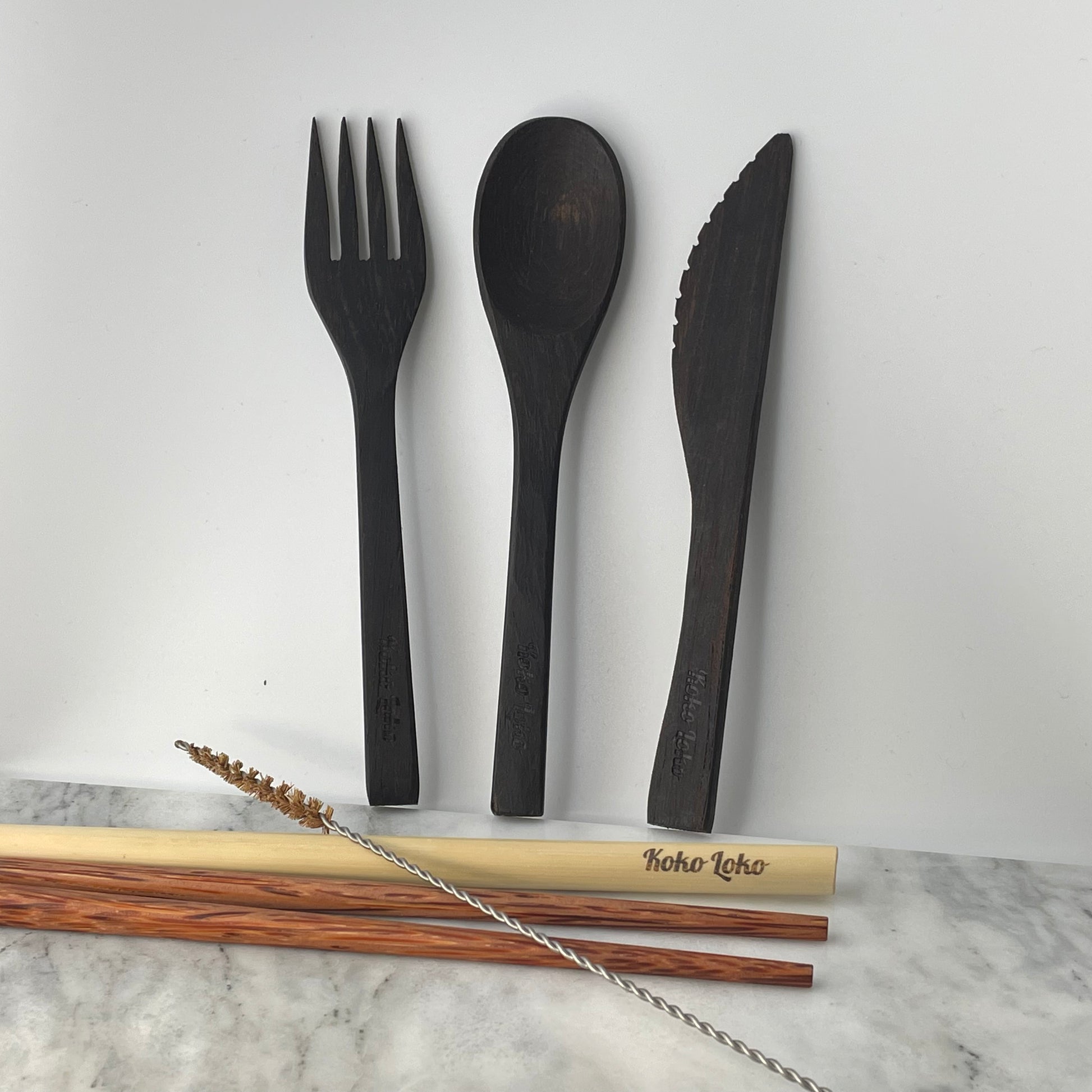 Nirvana Cutlery Collection handcrafted ebony wood fork, spoon and knife with Koko Loko engraving standing upright against a white wall above dark wood chopsticks, bamboo straw and cleaner brush on a marble surface, eco-friendly sustainable cutlery set by Koko Loko

