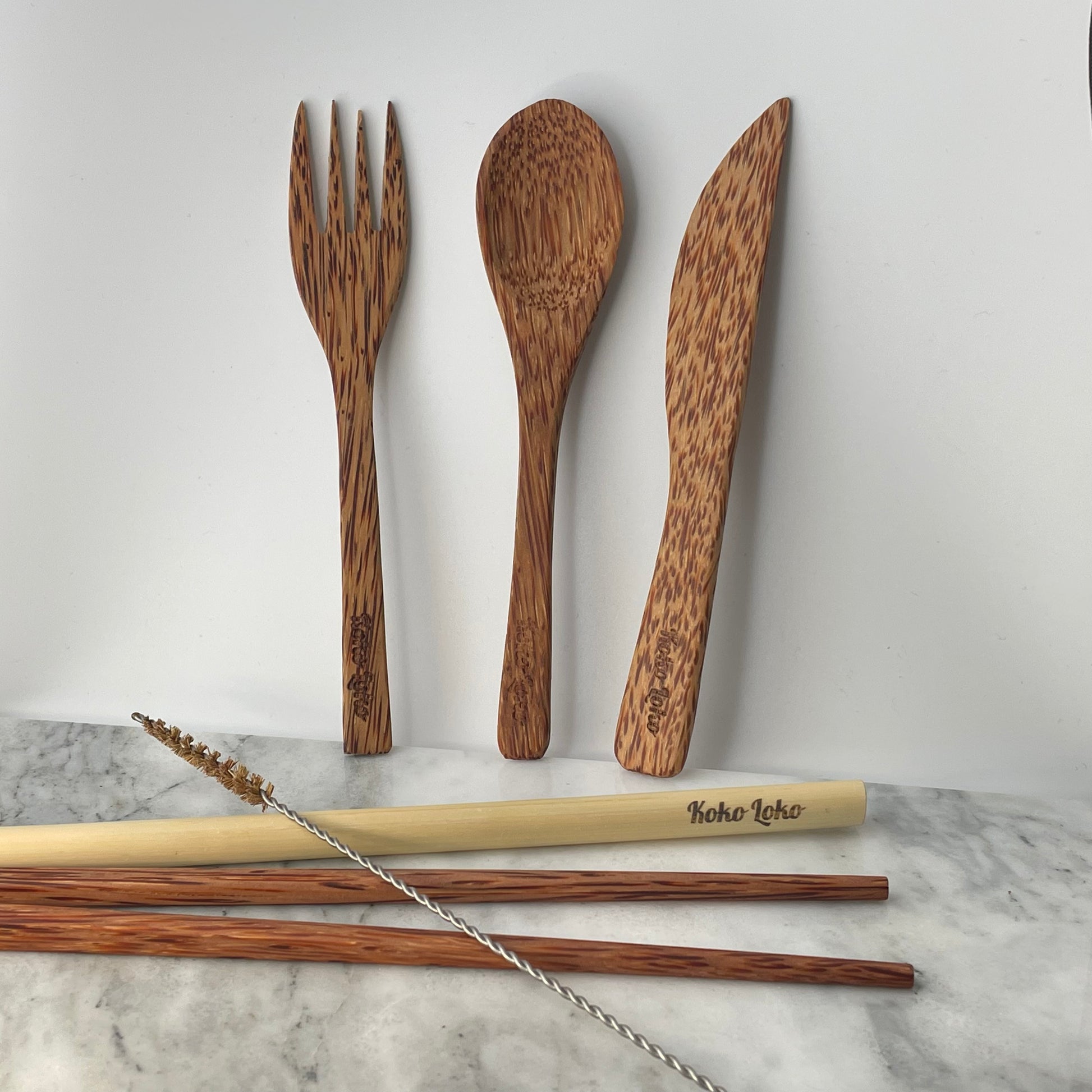 Earth Cutlery Collection handcrafted coconut wood fork, spoon and knife with Koko Loko engraving standing upright against a white wall above dark wood chopsticks, bamboo straw and cleaner brush on a marble surface, eco-friendly sustainable cutlery set by Koko Loko
